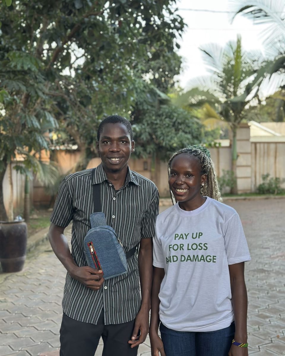 Geofrey (left, wearing a striped shirt and a small bag) & Clare (right, wearing a white 'PAY UP FOR LOSS AND DAMAGE' shirt) outdoors on a paved area with trees and a fence in the background.