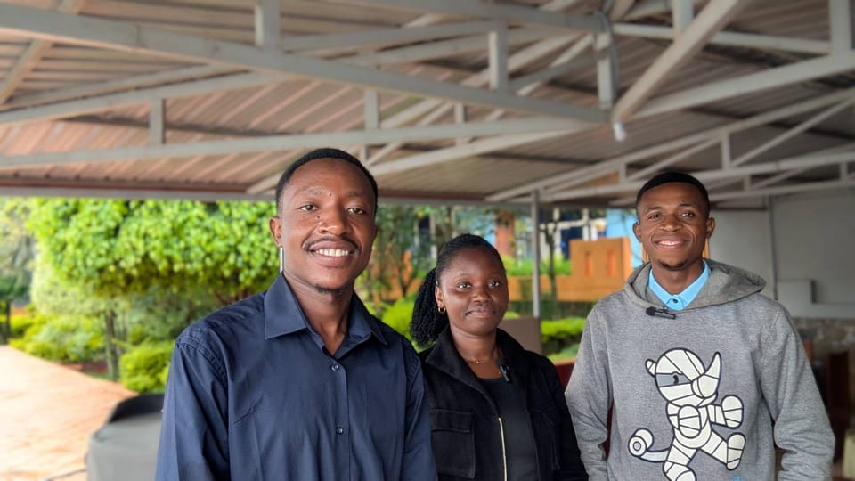 Three people standing under a metal-roofed shelter with greenery in the background.